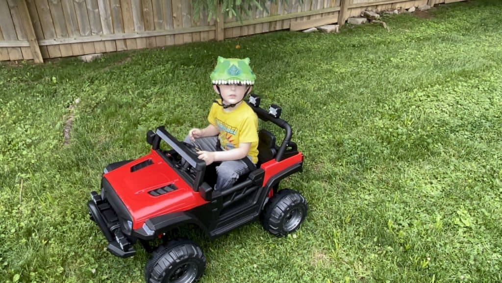 Kevin's son in a green dinosaur helmet driving a red ride-on jeep across the backyard grass